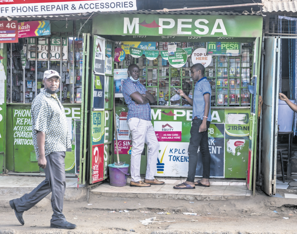 Mehrere Männer stehen vor einem kleinen grünen M-PESA-Shop in einer belebten Straße. Der Laden ist mit zahlreichen Schildern und Logos verschiedener Mobilfunk- und Finanzdienstleister bedeckt, darunter Safaricom, Telkom und Equity Bank. Ein Mann bedient Kunden am Schalter, während andere vor dem Geschäft warten oder vorbeigehen.