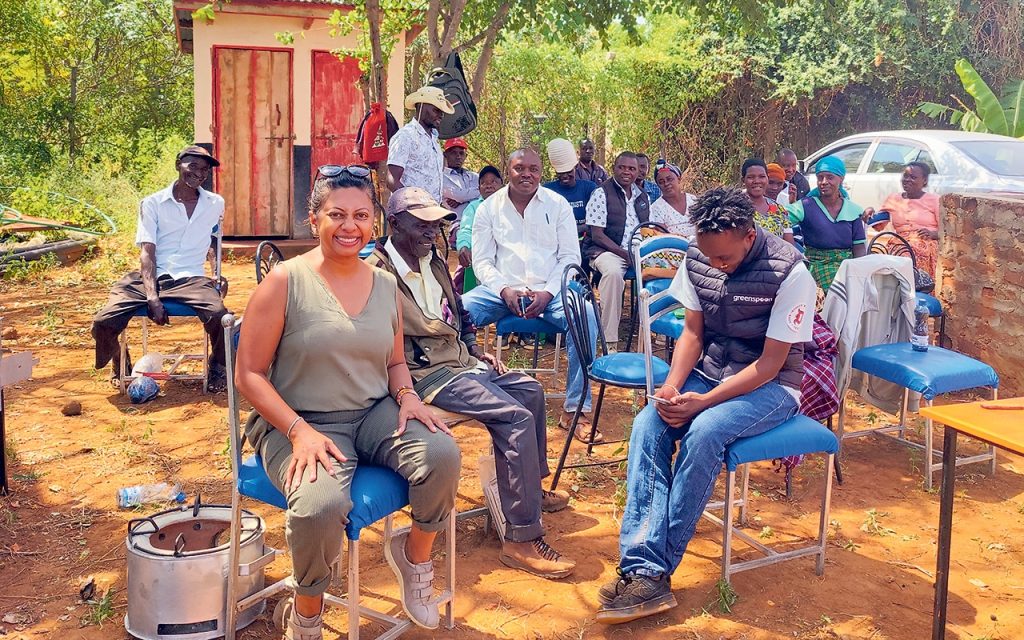 Eine Gruppe von Menschen sitzt im Freien unter Bäumen auf Stühlen in einer lockeren Gesprächsrunde. Eine Frau im Vordergrund lächelt in die Kamera, während andere Personen aufmerksam zuhören oder sich unterhalten. Im Hintergrund sind ein kleines Gebäude und ein Auto zu sehen.