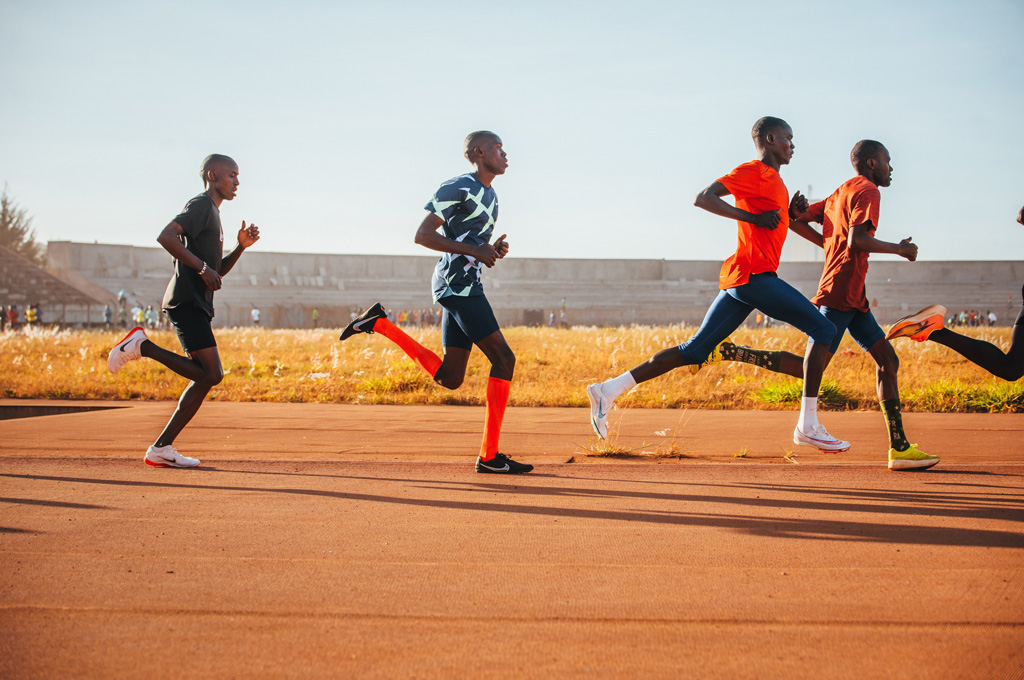 Fünf Läufer trainieren auf einer Laufbahn bei sonnigem Wetter, sie bewegen sich dynamisch von links nach rechts. Im Hintergrund sind ein Stadion und eine Wiese zu sehen