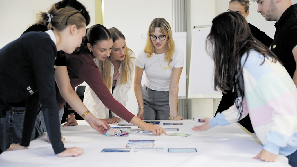 Eine Gruppe von Studierenden steht um einen Tisch und diskutiert gemeinsam.