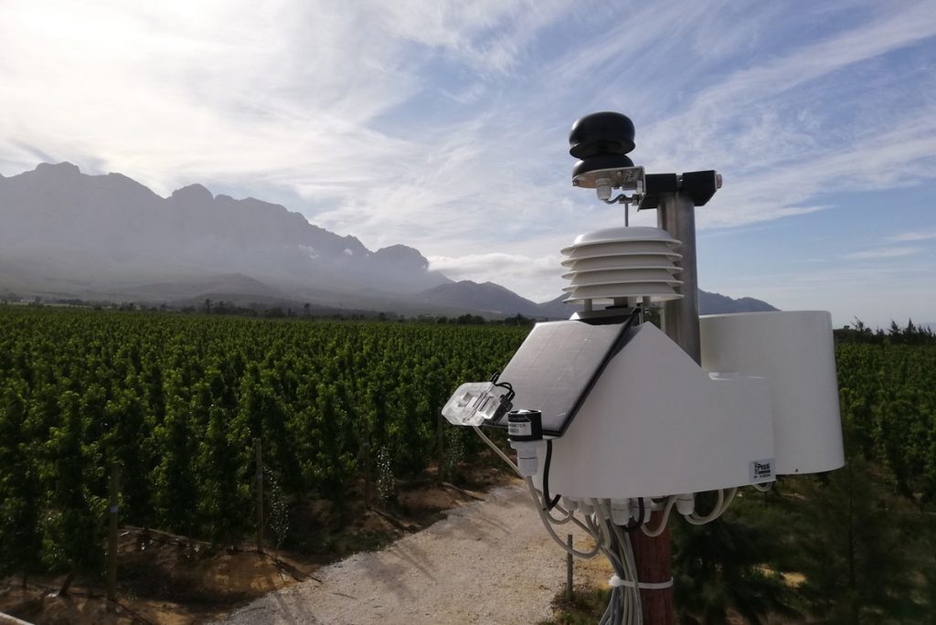 Eine Wetterstation mit Solarpanel und Sensoren steht am Rand eines grünen Feldes mit Bergen und Wolken im Hintergrund.