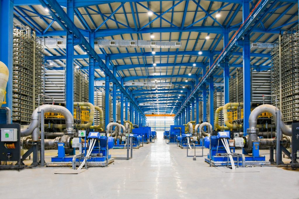 Innenansicht einer Entsalzungsanlage mit blauen Stahlträgern, großen Rohren, Pumpen und Filtereinheiten in einer hellen Industriehalle.