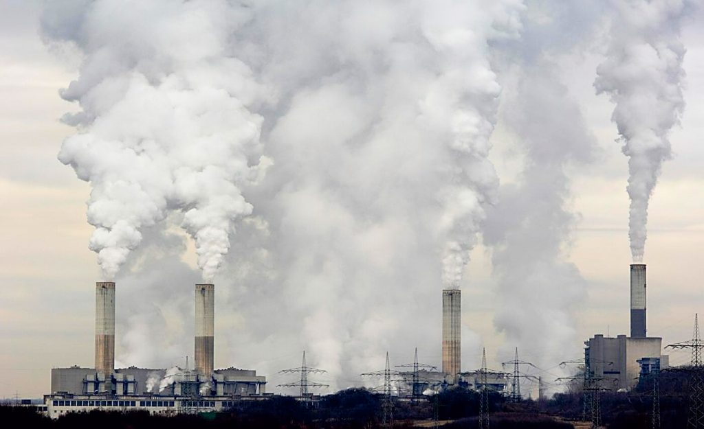 Großes Kohlekraftwerk mit mehreren Schornsteinen, aus denen dichte weiße Rauchwolken aufsteigen. Stromleitungen verlaufen im Vordergrund.