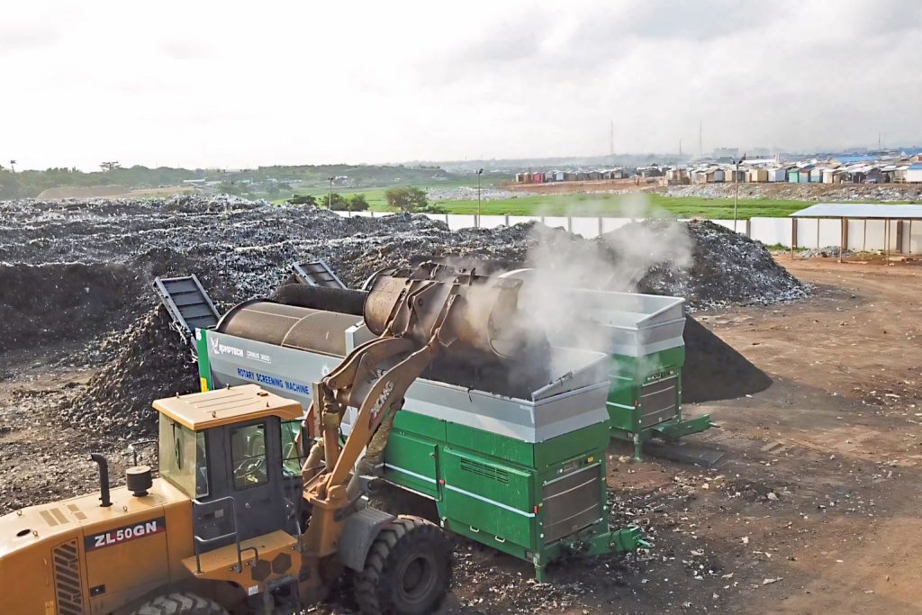 Ein Radlader lädt organisches Material in eine grüne Siebmaschine auf einer Kompost- oder Recyclinganlage, aus der Dampf aufsteigt.