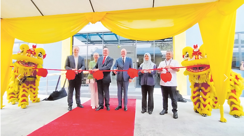 Sechs Personen schneiden gemeinsam ein rotes Band bei einer feierlichen Eröffnung unter einem gelb geschmückten Zelt. Zwei Löwentanzfiguren stehen links und rechts daneben.