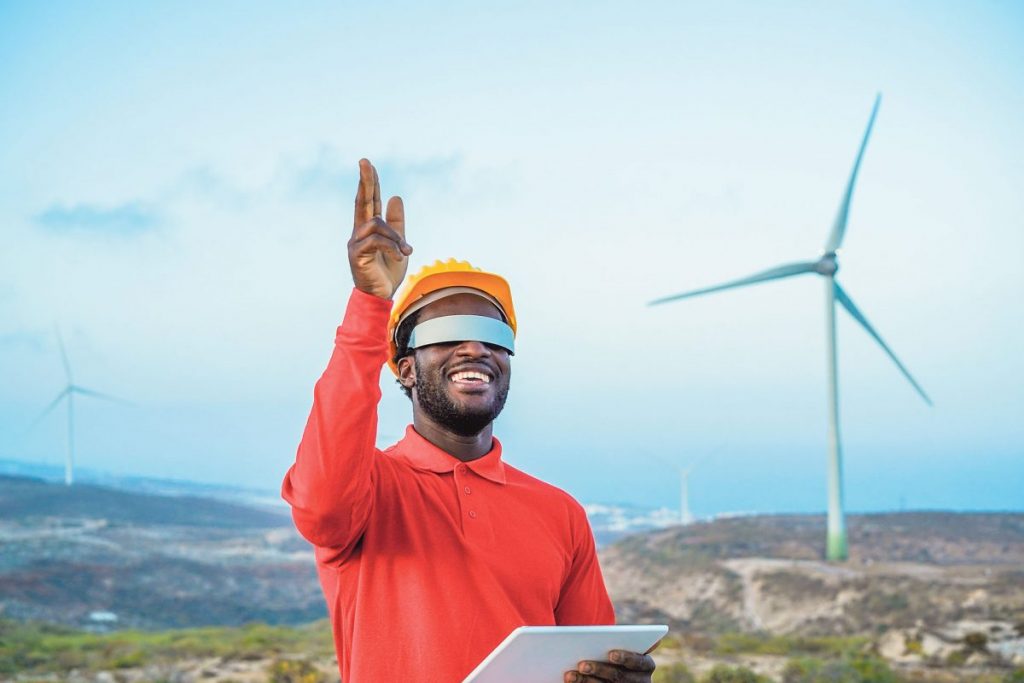 Ein Mann mit Schutzhelm und futuristischer AR/VR-Brille steht im Freien vor Windrädern, hält ein Tablet in der Hand und zeigt nach oben. Er lächelt und symbolisiert den Einsatz digitaler Technologien im Bereich erneuerbarer Energien und moderner Industrie.