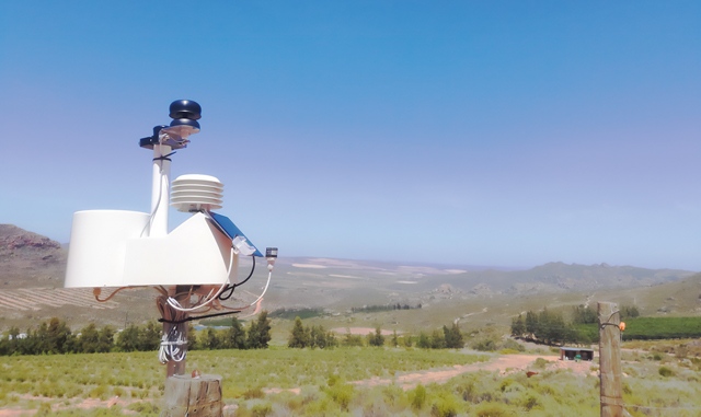Eine automatische Wetterstation mit Sensoren steht auf einem Pfosten in einer weiten, sonnigen Landschaft mit Feldern und Hügeln im Hintergrund.