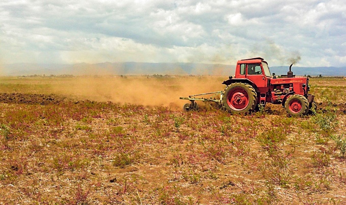 Ein roter Traktor pflügt ein trockenes Feld, während Staub aufwirbelt; im Hintergrund sind Berge und bewölkter Himmel zu sehen.