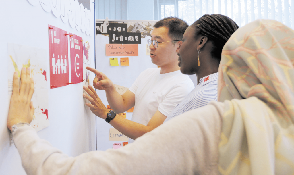 Drei Personen arbeiten gemeinsam an einer Pinnwand mit Karten zu den UN-Nachhaltigkeitszielen „No Poverty“ und „Reduced Inequalities“. Eine Person zeigt erklärend auf eine Karte.