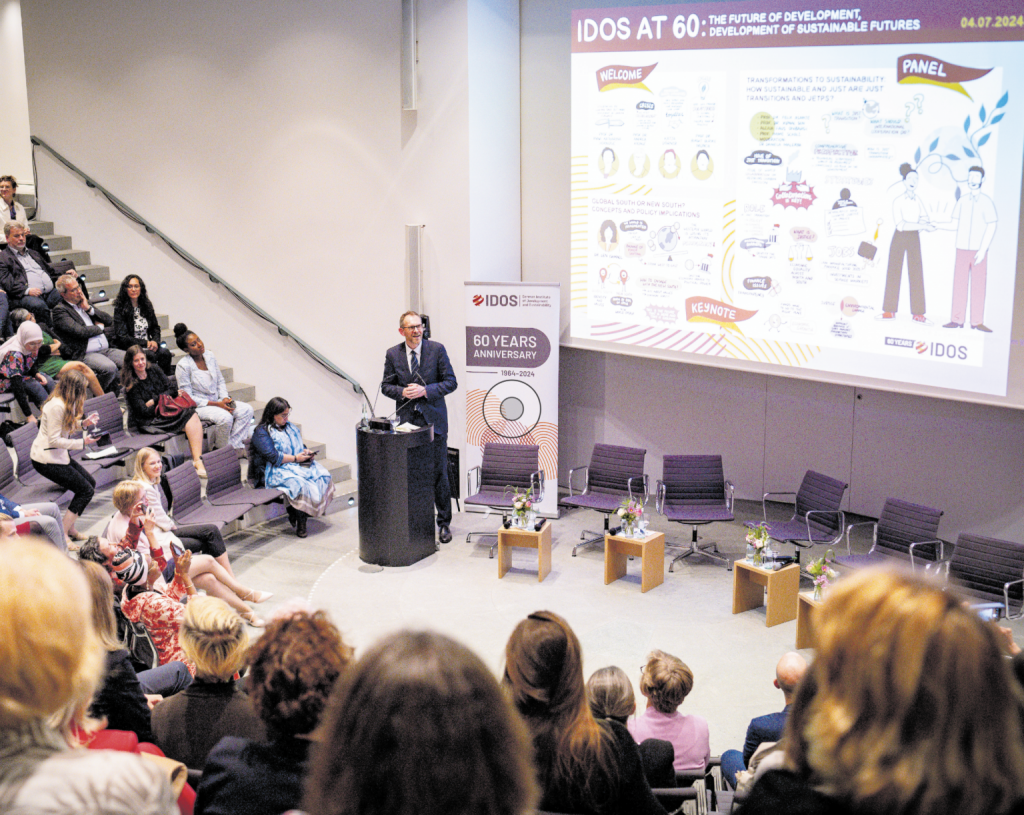 Ein Redner spricht am Pult vor einem Publikum in einem modernen Hörsaal. Auf der Leinwand hinter ihm steht „IDOS at 60: The Future of Development“. Mehrere Personen hören aufmerksam zu.
