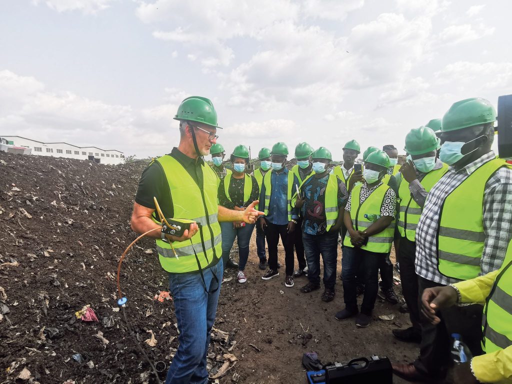 Eine Gruppe von Personen mit grünen Schutzhelmen und Warnwesten steht auf einer Deponie. Ein Mann erklärt technische Details und hält ein Messgerät in der Hand.