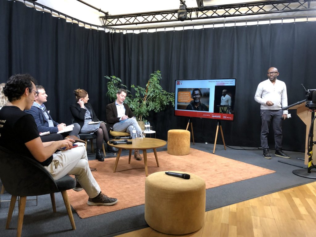 Mehrere Personen sitzen in einem Studio und hören einem Vortragenden zu, der neben einem Bildschirm mit Präsentationsfolien steht.