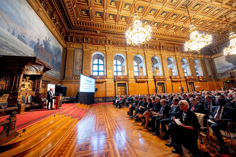 Zahlreiche Teilnehmende verfolgen eine Rede in einem prunkvollen, goldverzierten Saal mit großen Kronleuchtern.