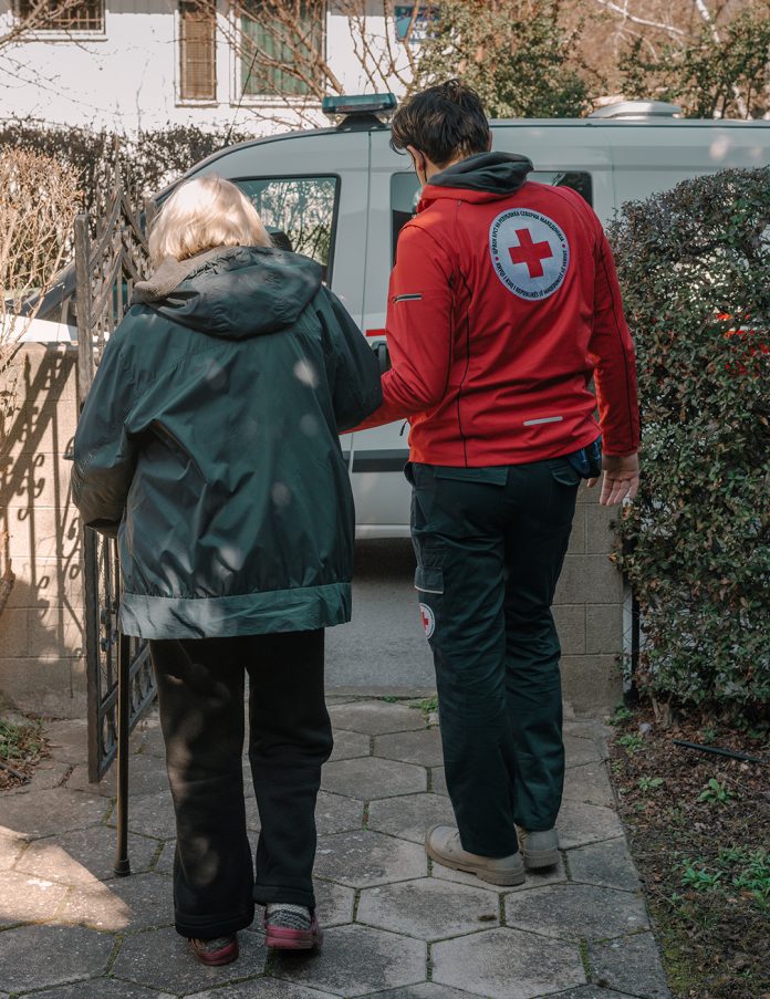 Eine Person in einer roten Jacke des Roten Kreuzes begleitet eine ältere Person beim Gehen durch einen Gartenweg.