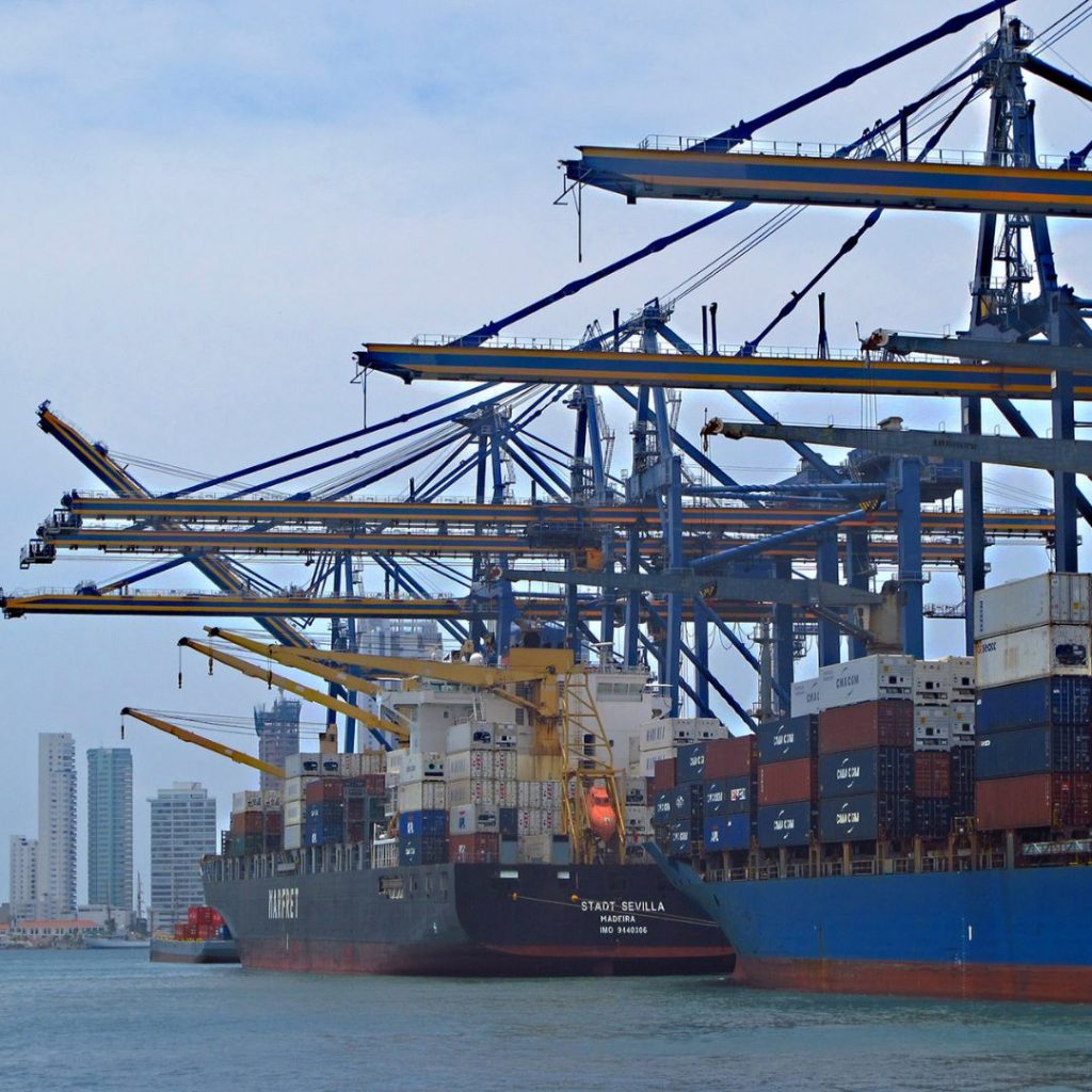 Containerhafen mit mehreren großen Kränen und Frachtschiffen, die Container be- und entladen. Im Hintergrund sind Hochhäuser der Stadt Cartagena zu sehen.