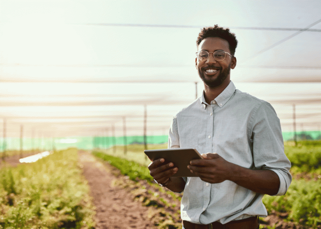 Ein Mann steht lächelnd in einem modernen landwirtschaftlichen Betrieb und hält ein Tablet in den Händen. Im Hintergrund sind bepflanzte Reihen unter einer schützenden Überdachung zu sehen.