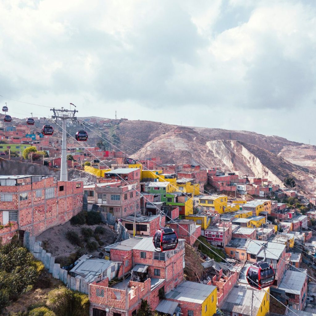 Bunte Häuser in einem hügeligen Stadtviertel mit einer modernen Seilbahn, die über die Dächer hinwegführt, vor einer bergigen Landschaft und bewölktem Himmel.