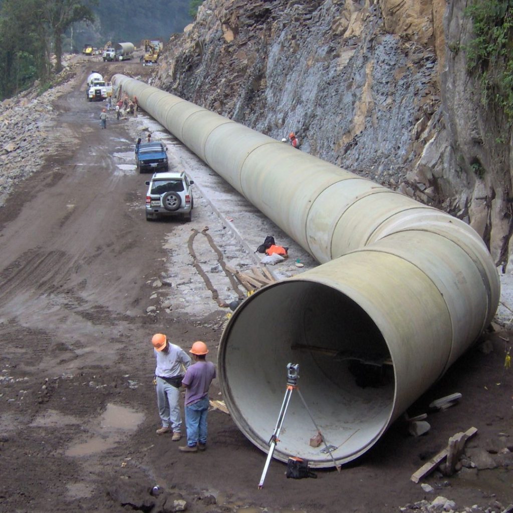Große Rohrleitungen werden entlang einer Gebirgsstraße verlegt. Arbeiter mit Schutzhelmen und Fahrzeuge sind auf der Baustelle zu sehen.