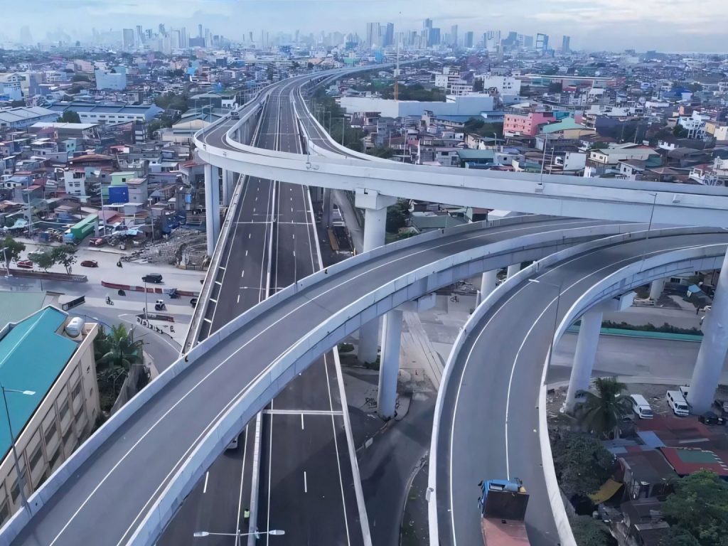 Luftaufnahme eines modernen Autobahnkreuzes mit mehreren Ebenen über dicht bebautem Stadtgebiet.