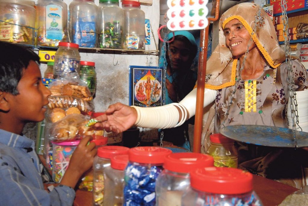 Frau in einem kleinen Laden reicht einem Jungen Ware über den Tresen, beide lächeln, umgeben von Gläsern mit Süßigkeiten und Keksen.